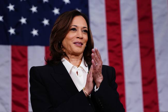 Kamala Harris portando un saco negro con camisa blanca y de fondo la bandera de los EE. UU.